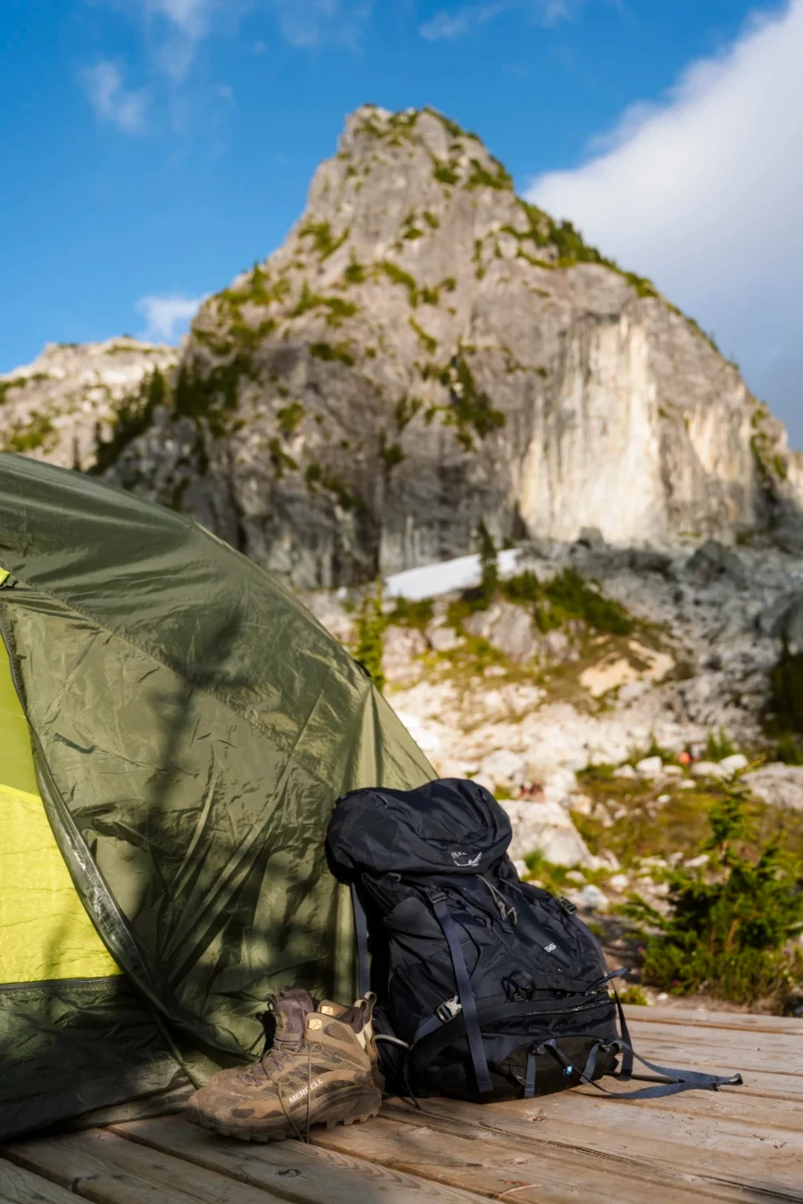 A pair of hiking boots and a backpack sit on a a wooden platform outside a green tent set up at the campsite at Watersprite Lake