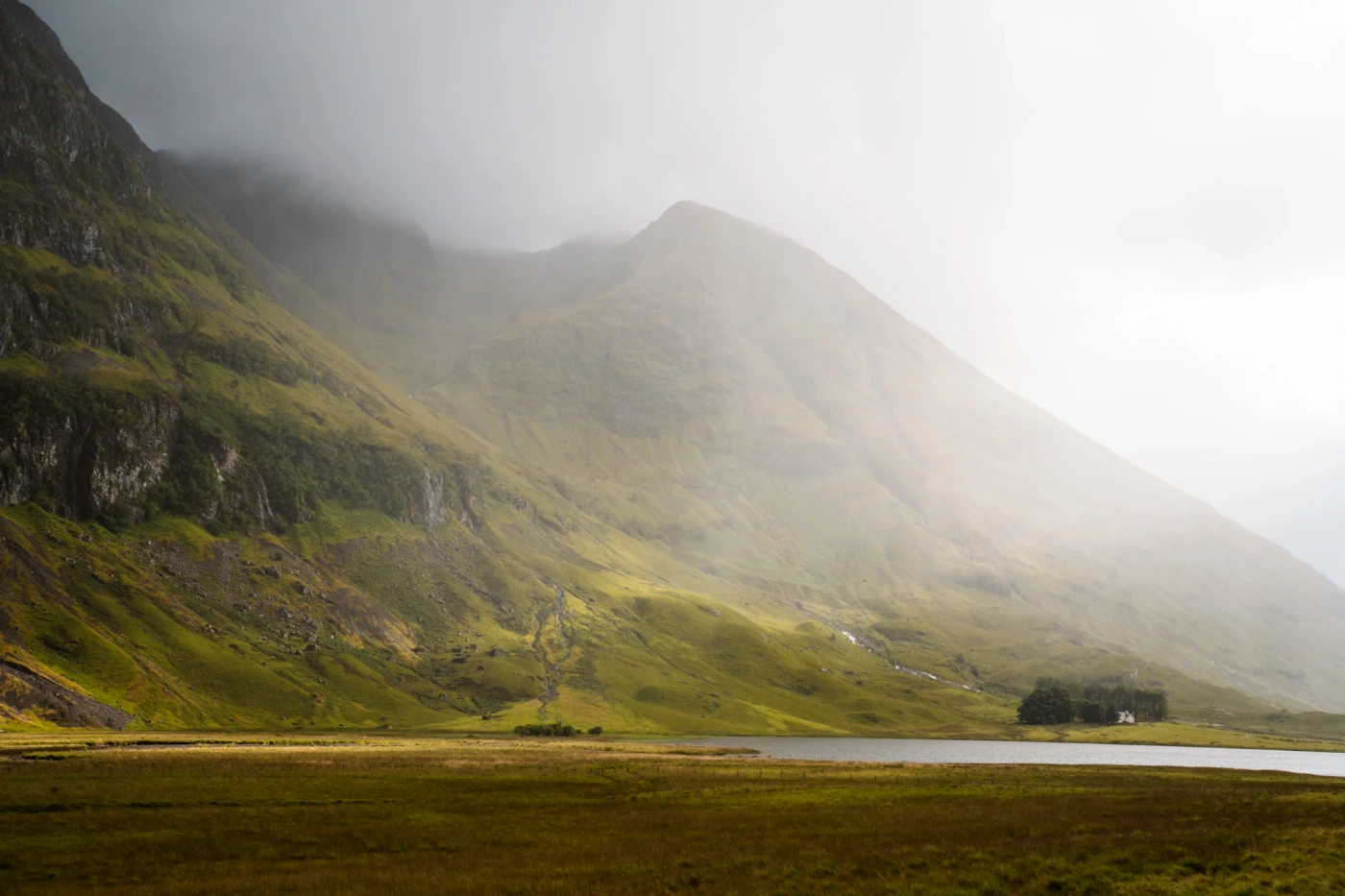 Mist rolls over a calm loch at the base of a tall grassy mountain in Glencoe, Scotland