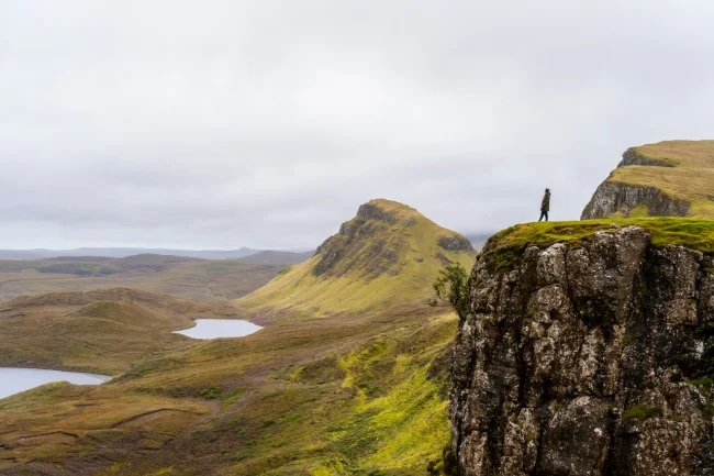 A man standing on the end of a large grass covered cliff with a wide grassy valley stretching out beneath him