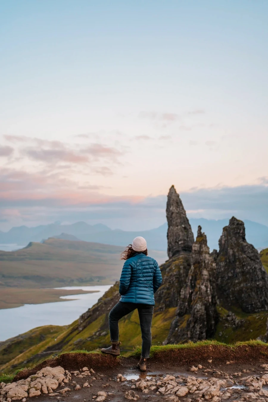 A woman standing on the wedge of a cliff with the iconic stone pillars of the Old Man of Storr in the distance