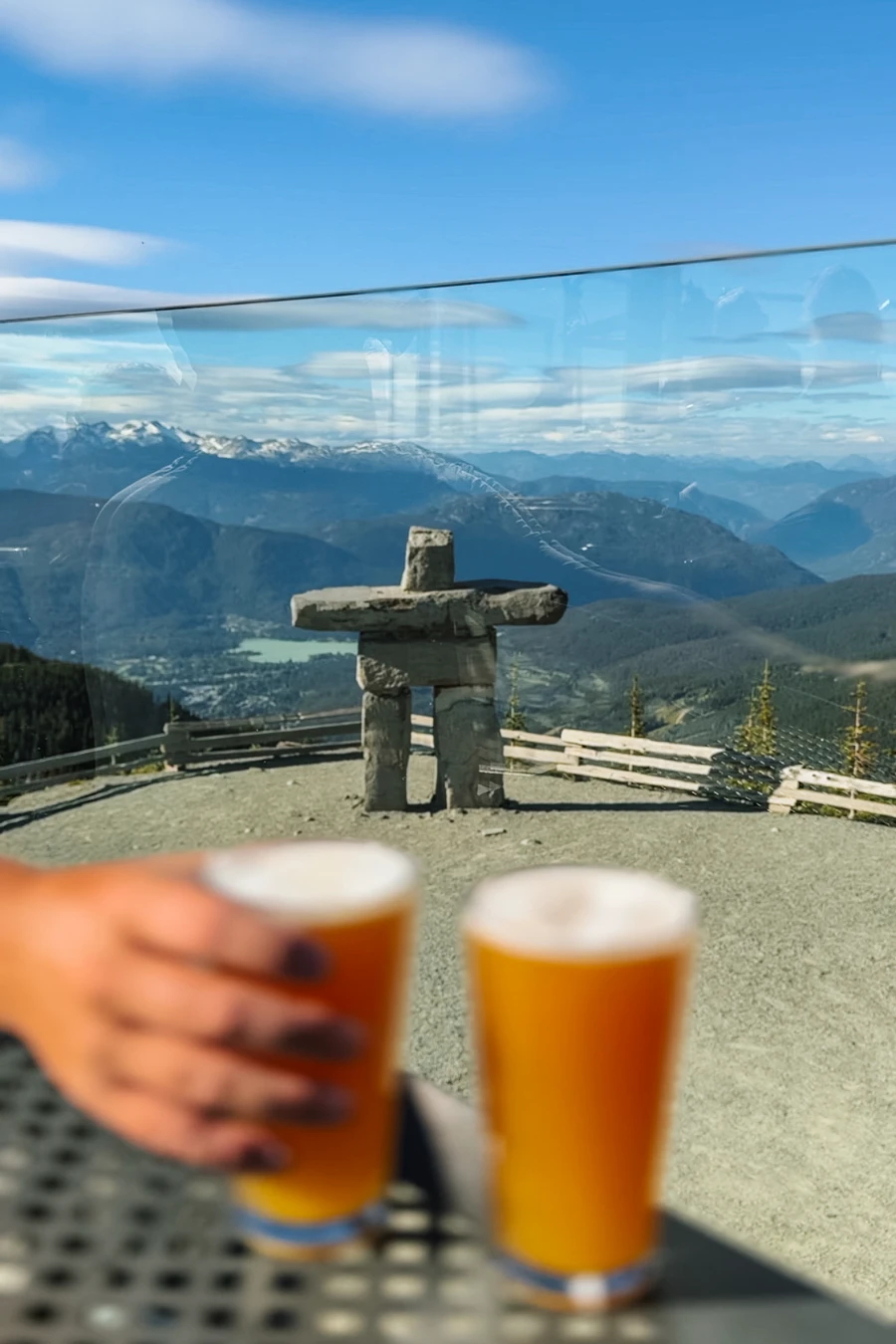 A hand hold one of two pints of beer in front of a lookout at the top of Whistler Mountain