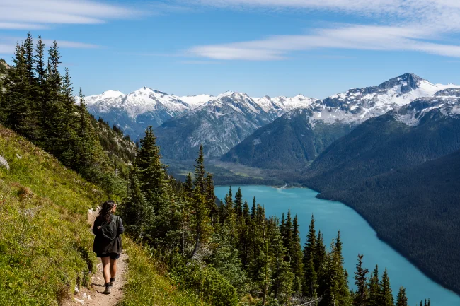 A woman walks along a trail in the Whistler alpine looking out at the snow-capped peaks that tower over Cheakamus Lake