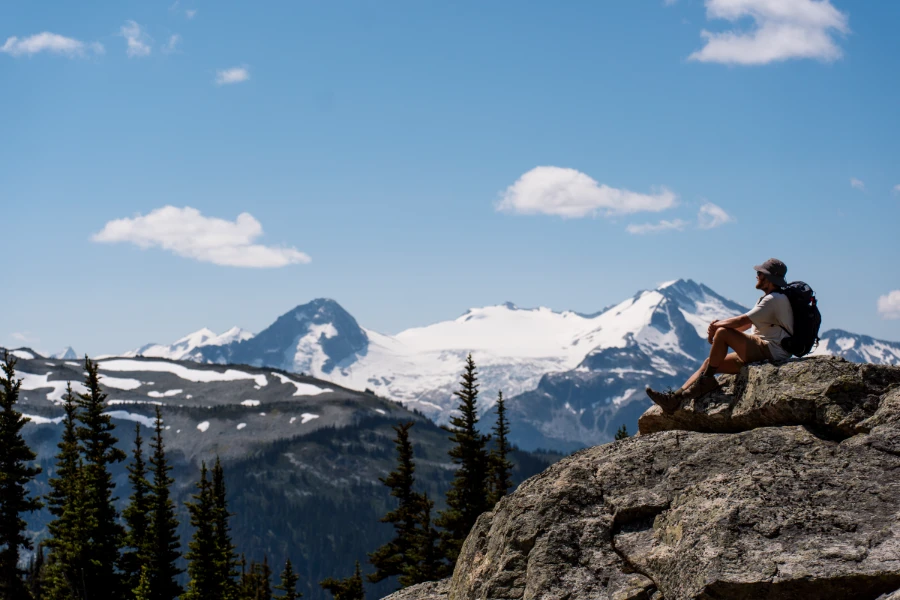 A man with a hiking backpack sitting on a boulder looking out to distant snow-capped mountains