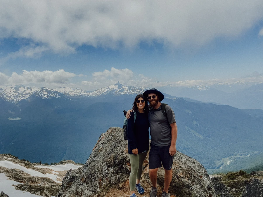 A man and woman stand arm in arm at the peak of Whistler Mountain with the distinctive shape of Black Tusk in the distance