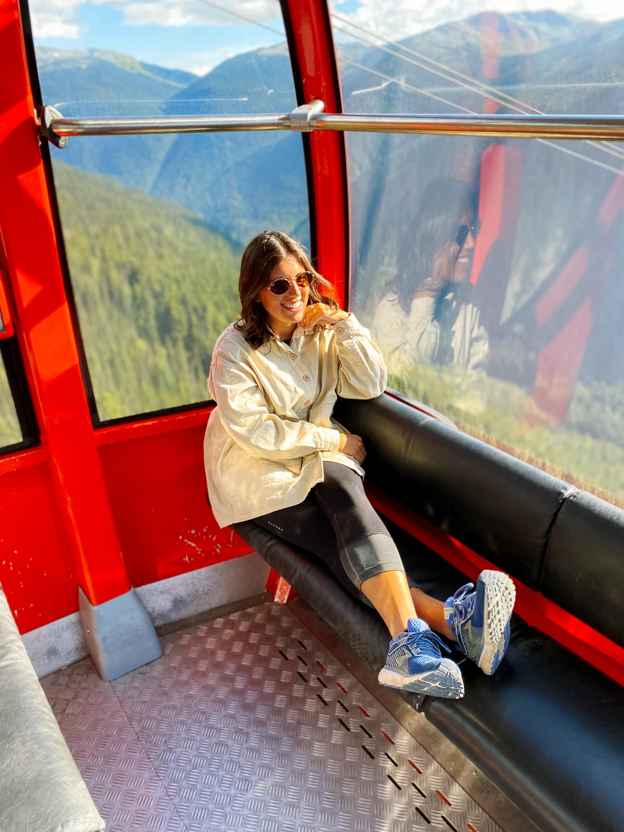 A woman sitting in a red Gondola with her legs up on the seath and a massive tree covered valley below