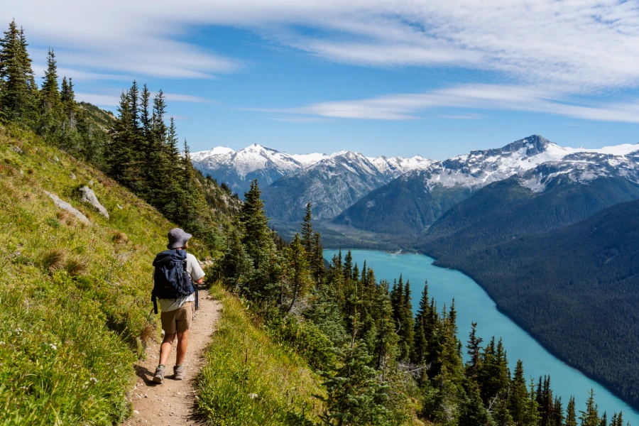 Man walking down a thing dirt path in the alpine with a large blue lake in the valley below to his side