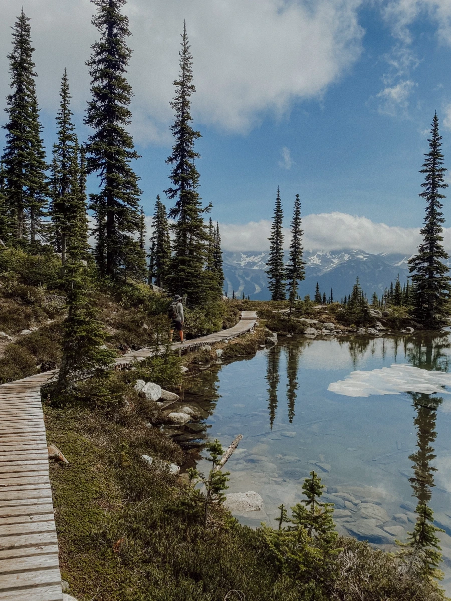 A wooden boardwalk borders a calm alpine lake with more mountains visible in the distance