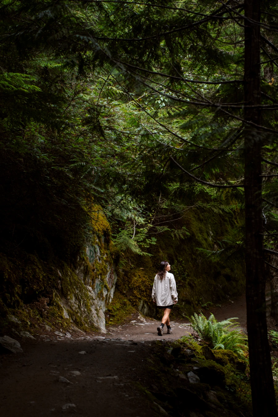 A woman walks along a flat trail that passes through a lush forest and next to a mossy cliff